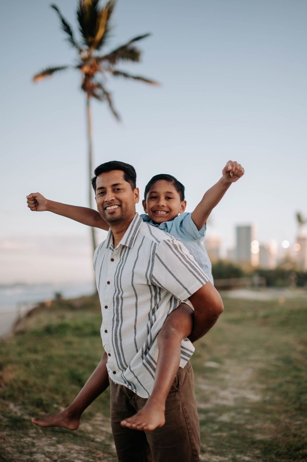 A joyful scene of play and bonding along a coastal path with palm trees and city buildings in the distance at sunset.