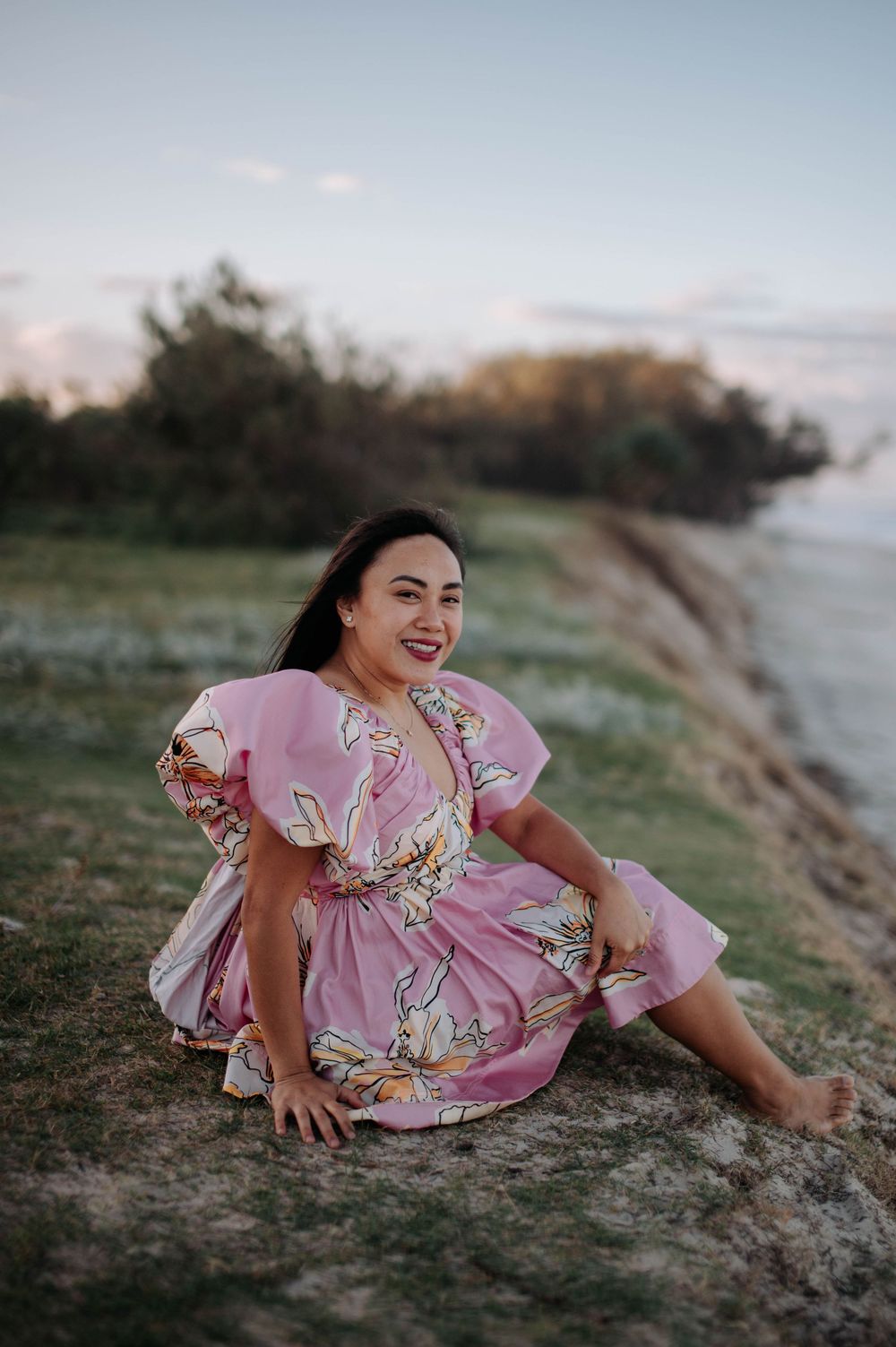 A woman wearing a pink floral dress sits on a grassy cliff overlooking the ocean at sunset.