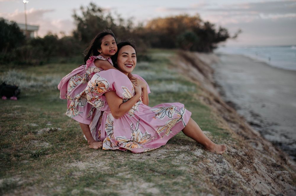 Two people in pink floral dresses sit together on a grassy cliff overlooking a beach at sunset.