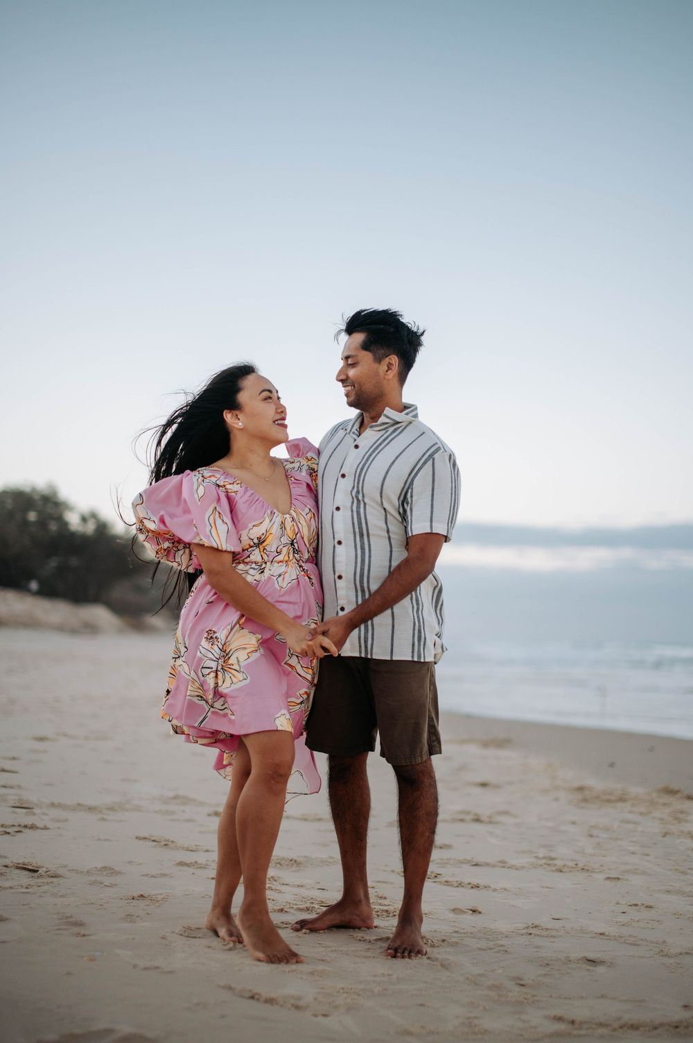 A happy couple in casual beach attire share a joyful moment together on a sandy shoreline at sunset.