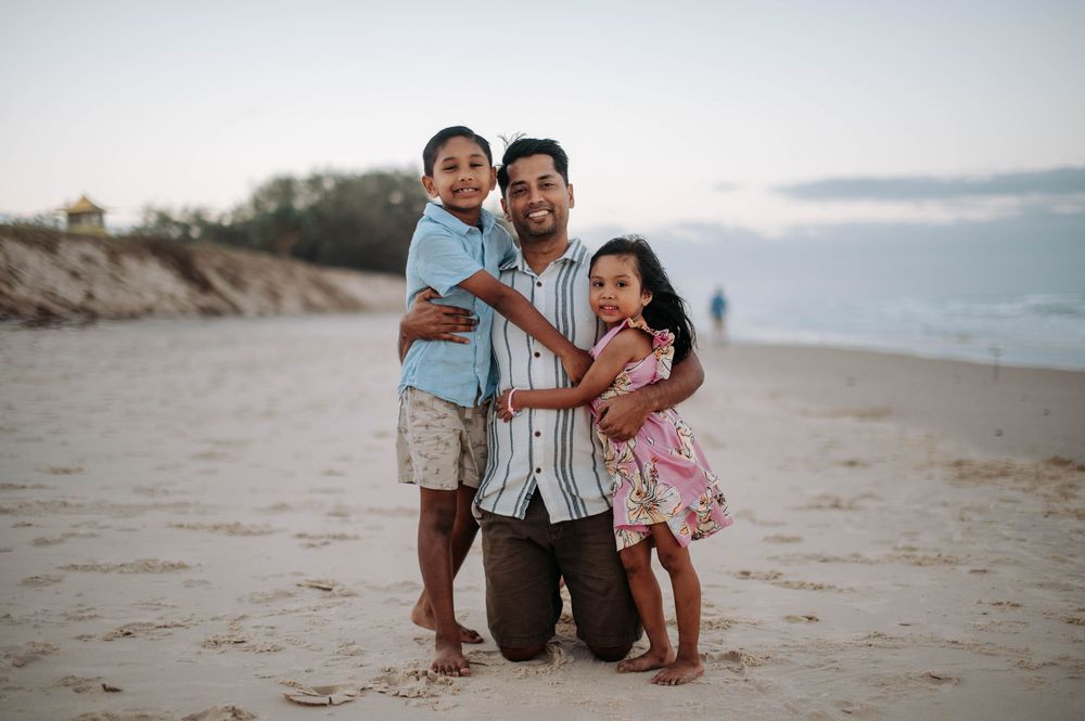 A family embraces together on a sandy beach at sunset with ocean waves and dunes in the background.