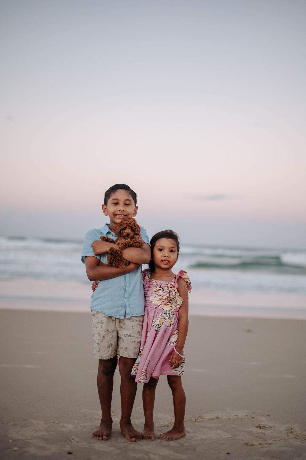 Two children standing barefoot on a beach at sunset holding a small reddish puppy.