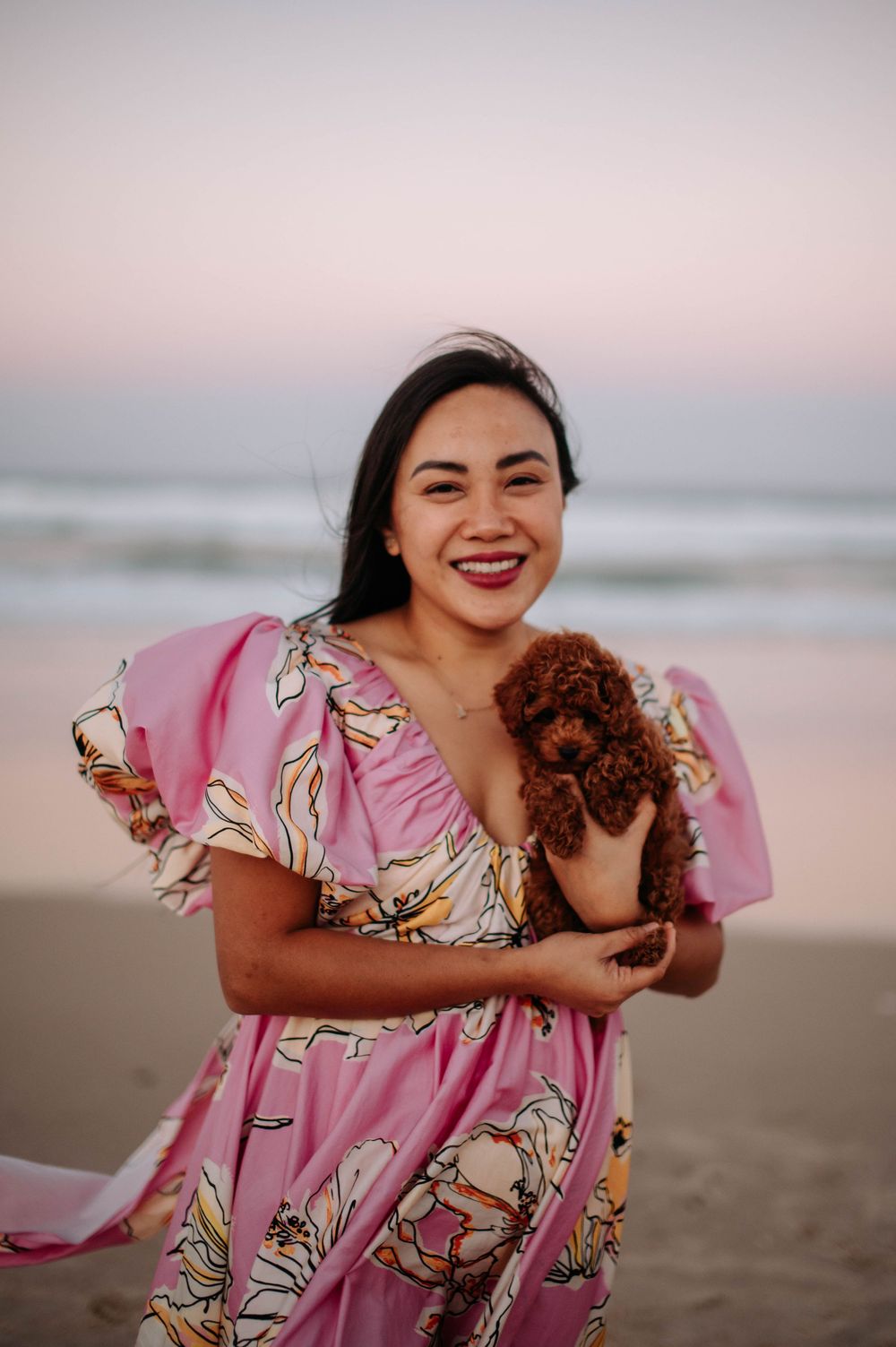 A woman in a pink floral dress holds a small brown puppy while smiling on the beach at sunset.