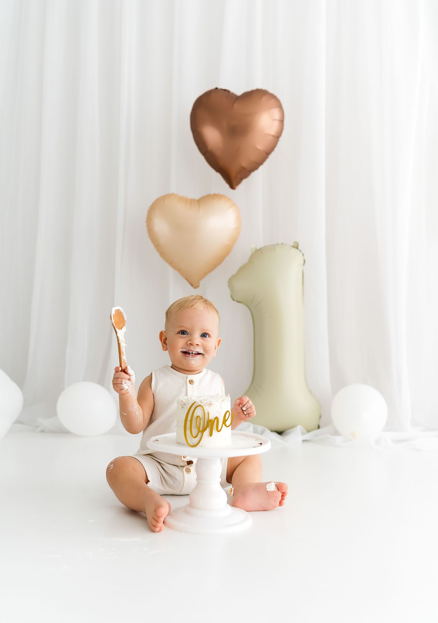 First birthday celebration setup with rose gold and cream heart balloons, number one balloon, and smiling baby holding spoon.