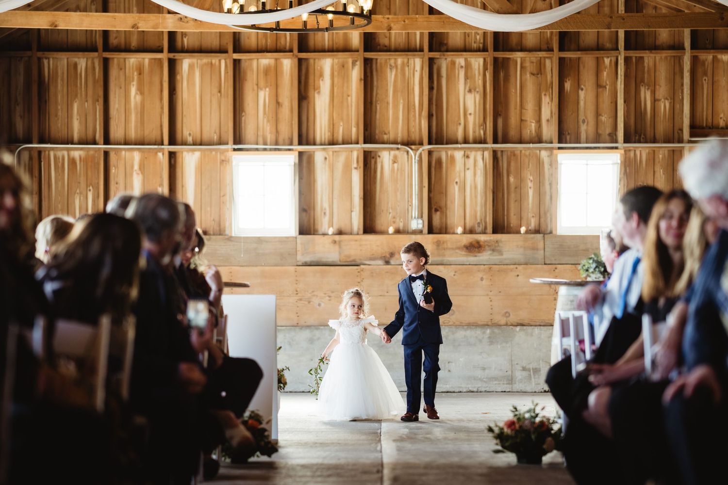 Wedding ceremony takes place inside rustic wooden barn with guests seated on both sides of the aisle.