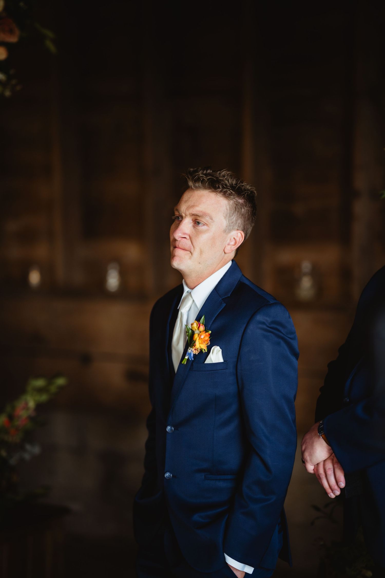 A person wearing a navy blue suit with an orange boutonniere poses in a dimly lit interior space.