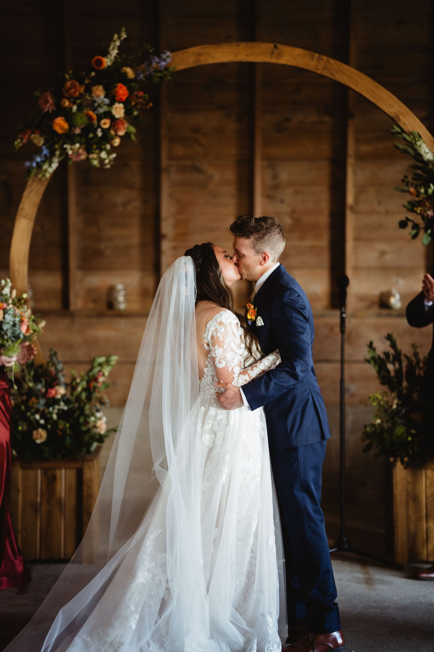 Elegant wedding ceremony with couple sharing a kiss under a wooden arch decorated with florals in a rustic barn setting.
