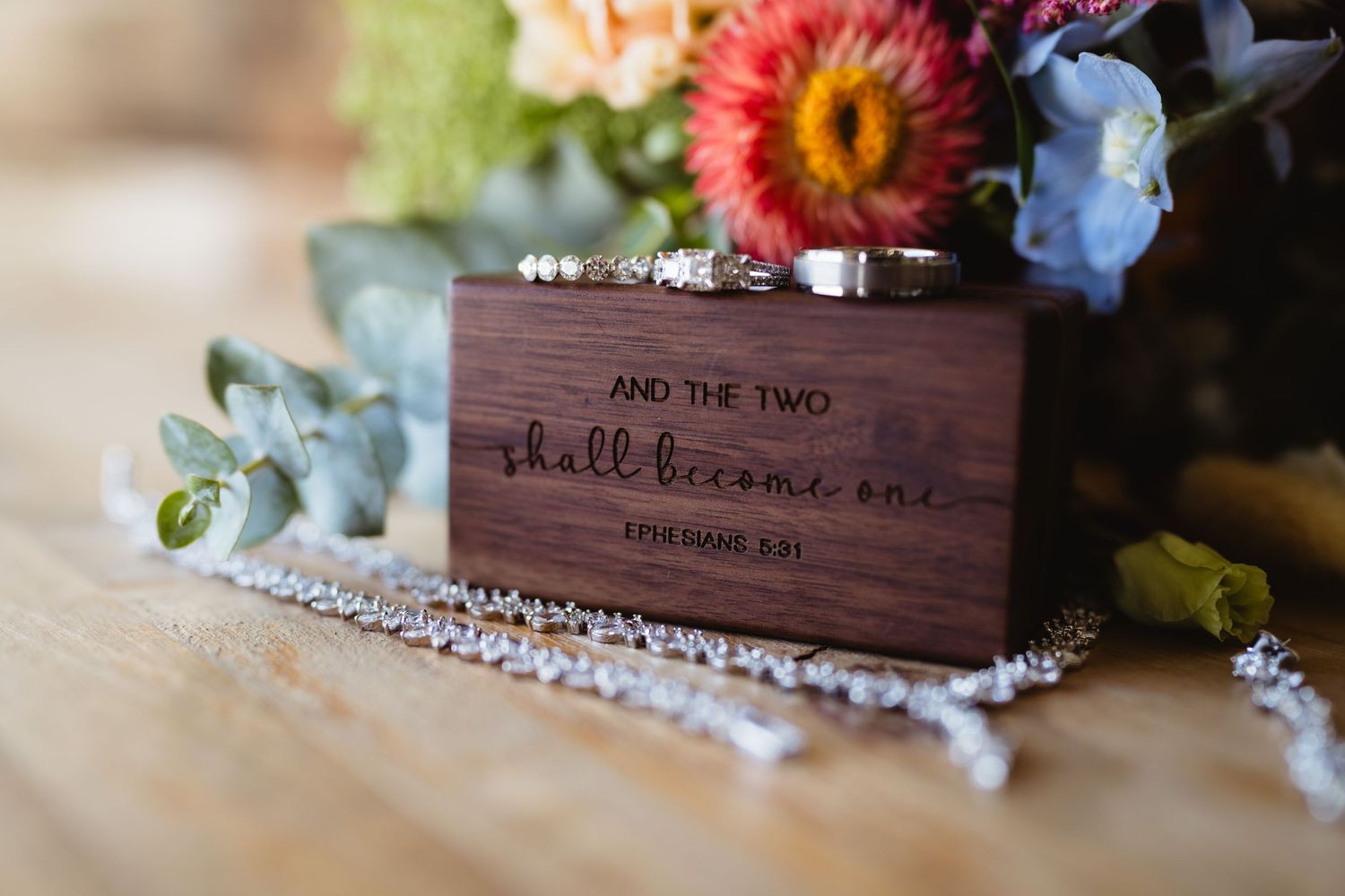 Wedding rings displayed on wooden box with scripture verse and floral decor against blurred background.