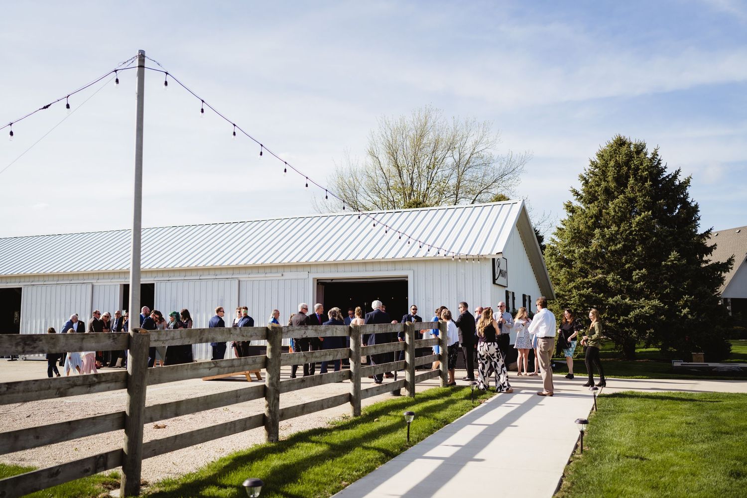 People gather outside a white barn venue with string lights and wooden fencing during an outdoor event celebration.