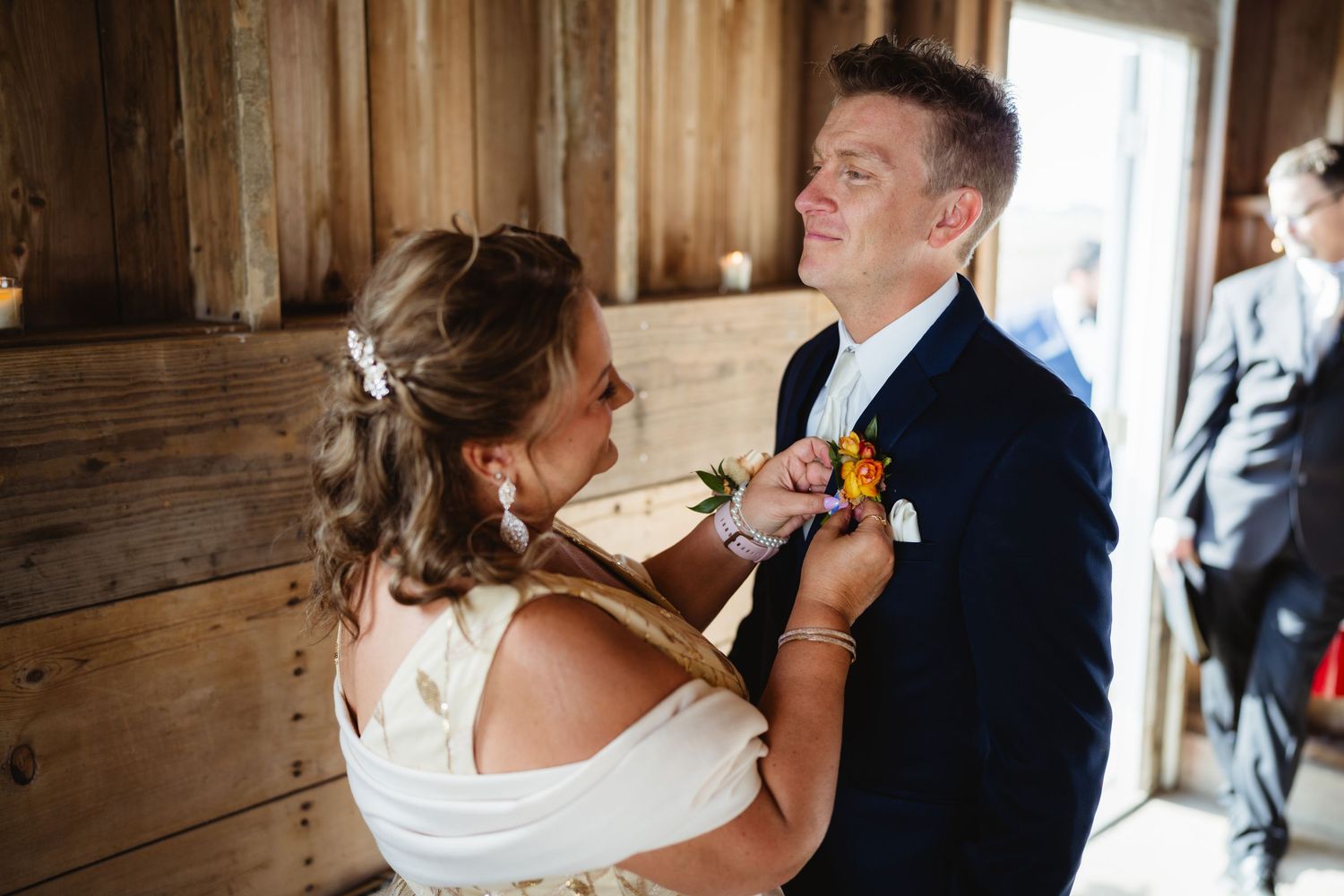 A couple shares an intimate moment during a wedding as they help each other with boutonniere in rustic wooden setting.