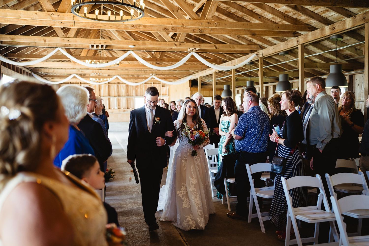 Wedding ceremony in a rustic barn venue with exposed wooden beams and white decorative lighting.