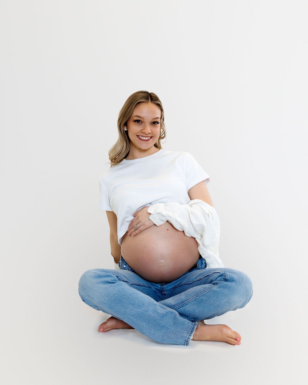 A pregnant person in jeans and white shirt sits cross-legged in three similar poses against a white backdrop.