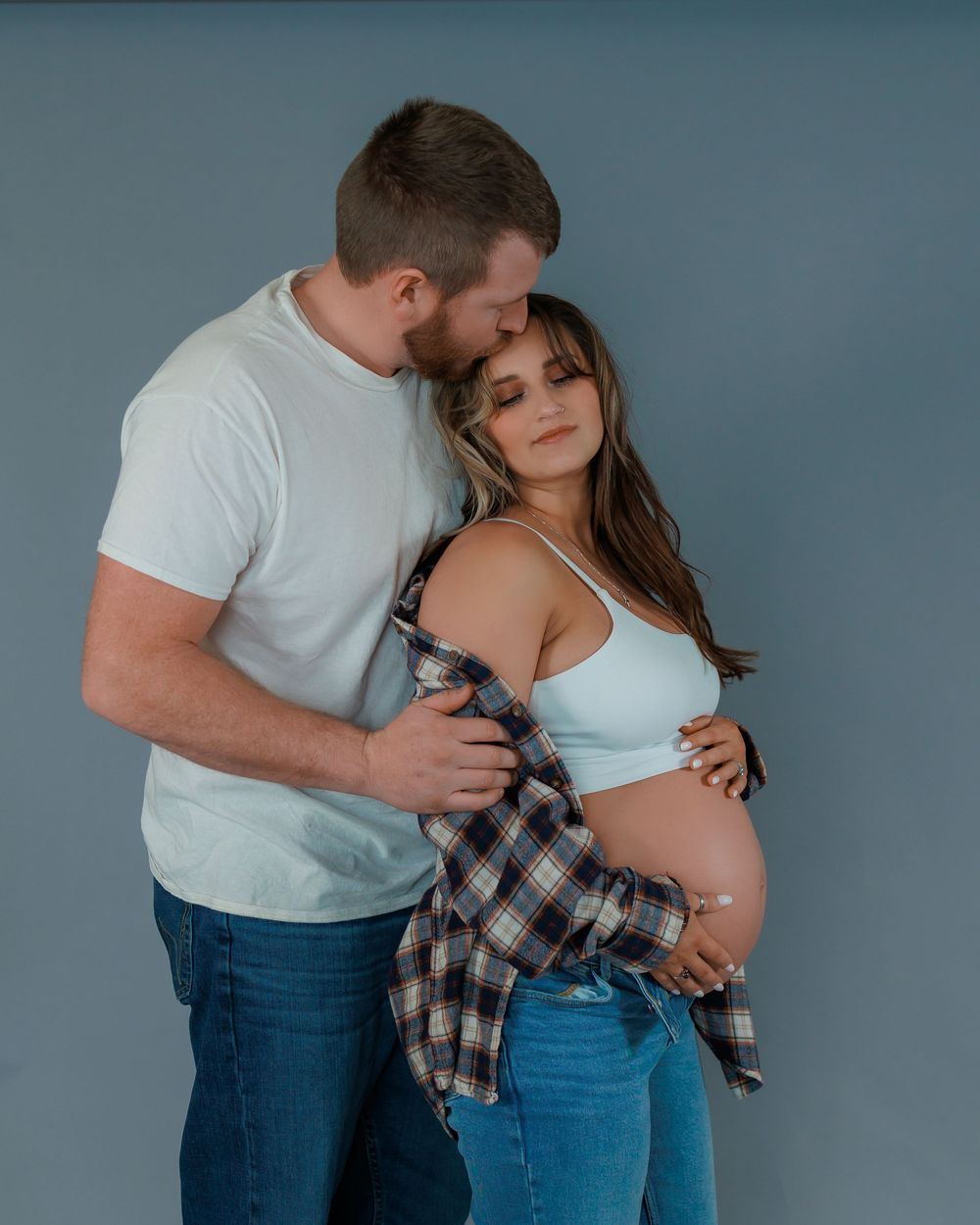 Intimate maternity photo of couple embracing against blue background, woman wearing plaid shirt and white top.