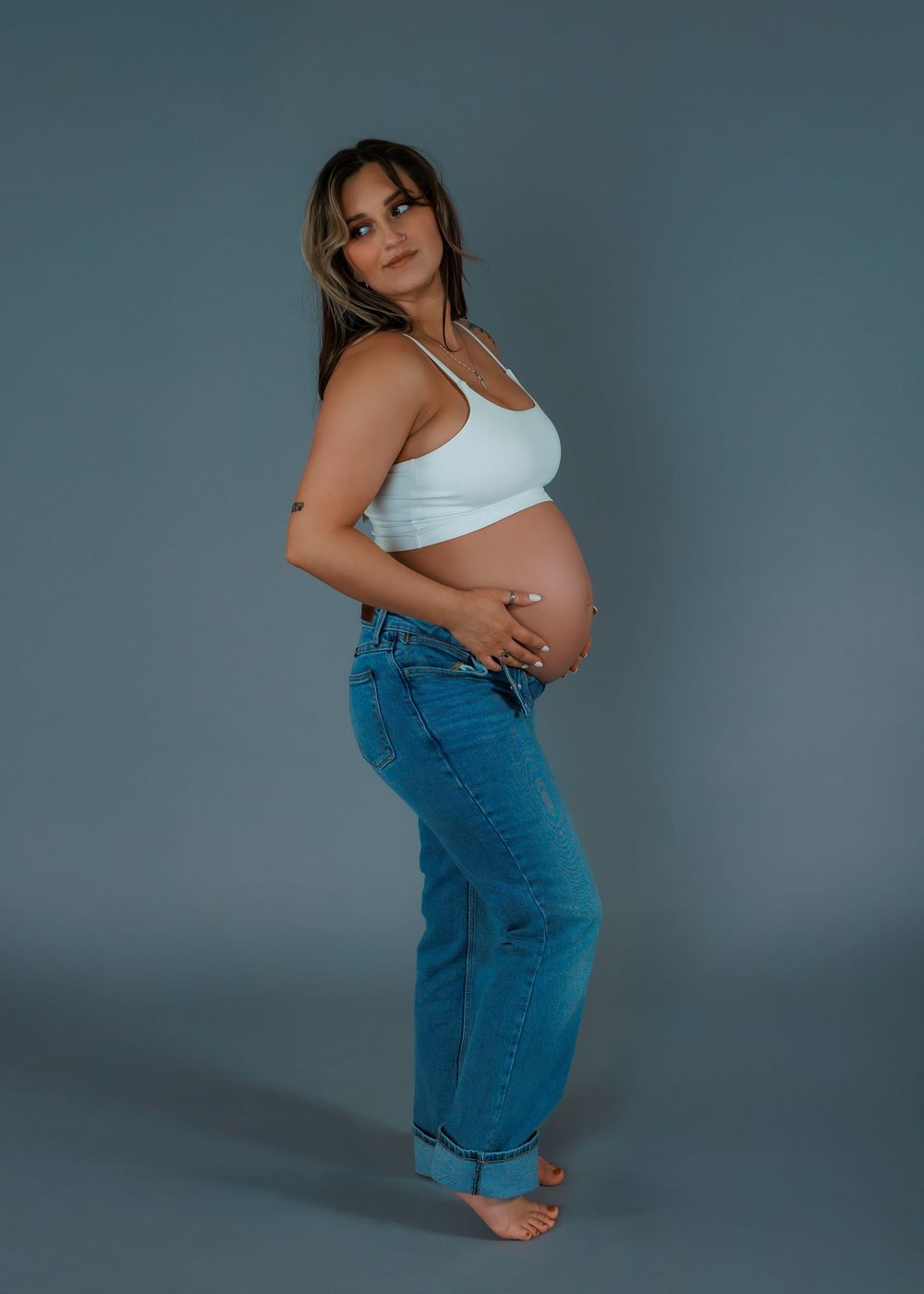 Side view of pregnant woman in white crop top and rolled jeans showing baby bump against blue backdrop.