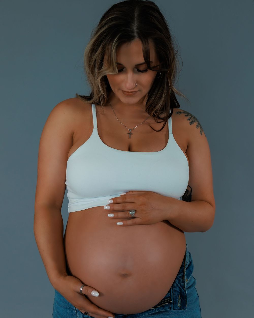 A pregnant woman in a white tank top and jeans cradles her baby bump against a dark blue background.