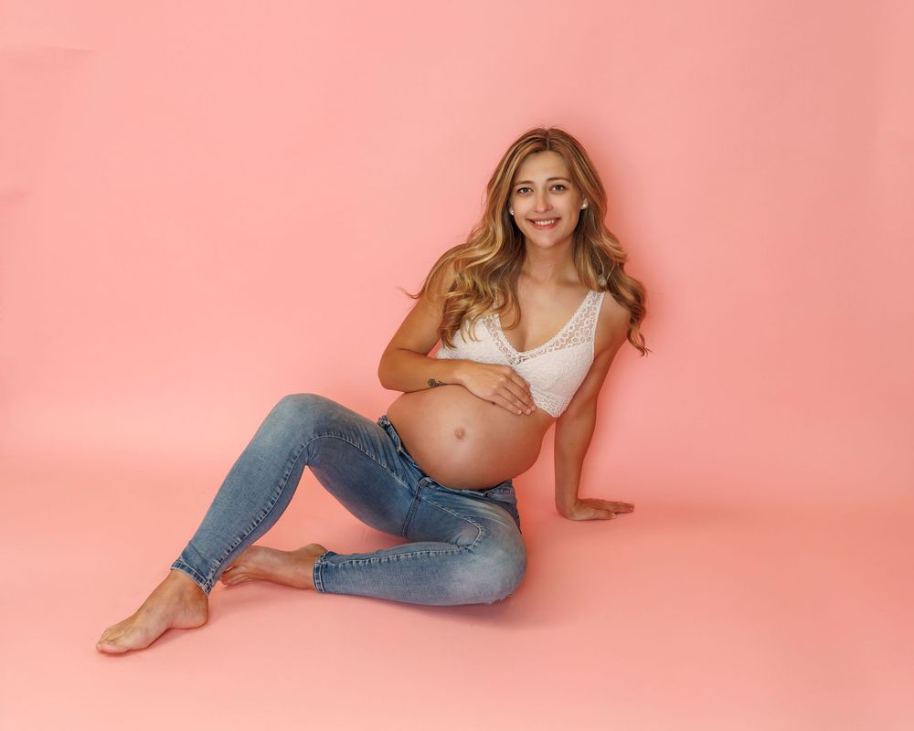 Expectant mother in white crop top and distressed jeans poses on pink studio backdrop.