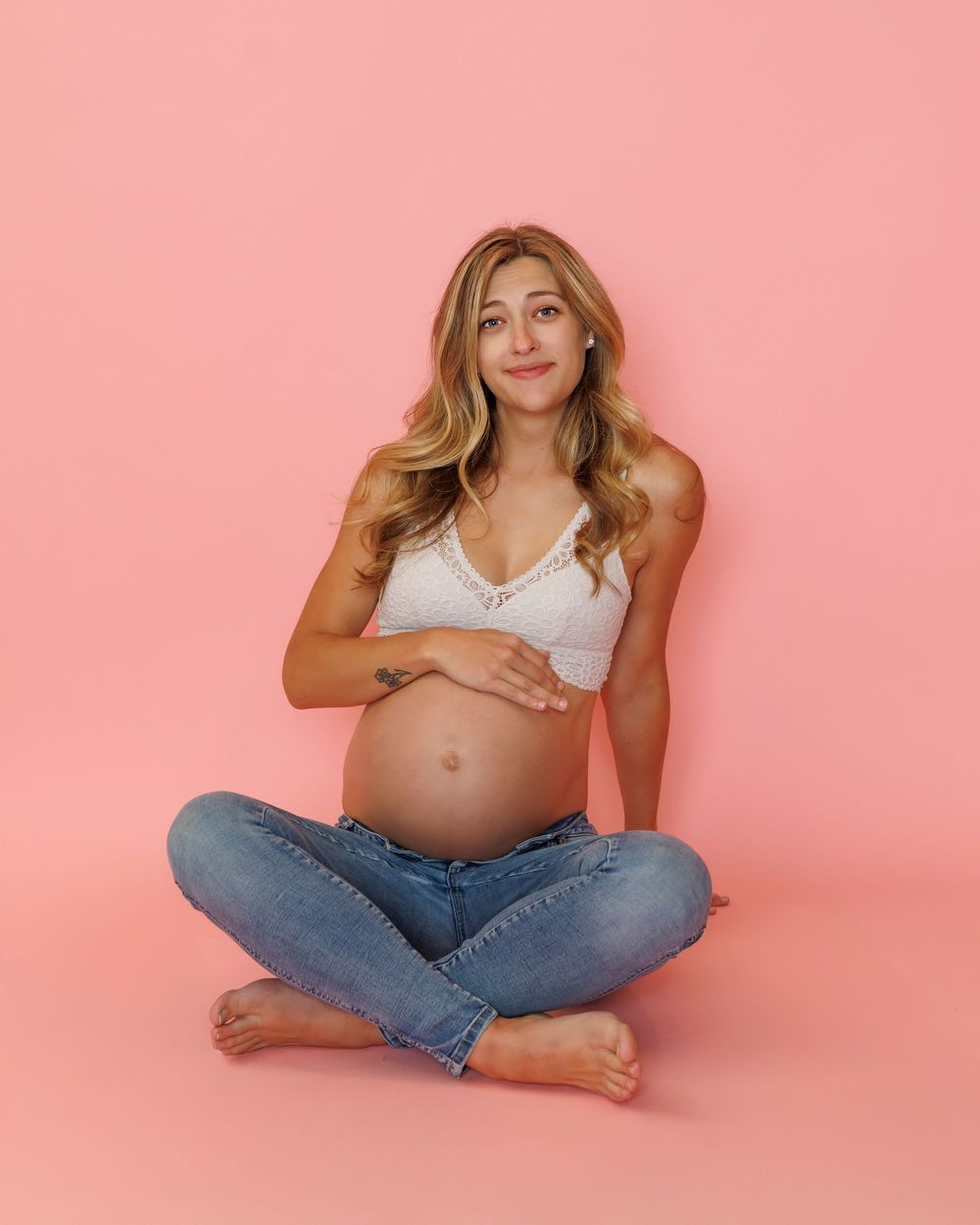 A pregnant woman in blue jeans and white crop top sits cross-legged on pink background in maternity photo.