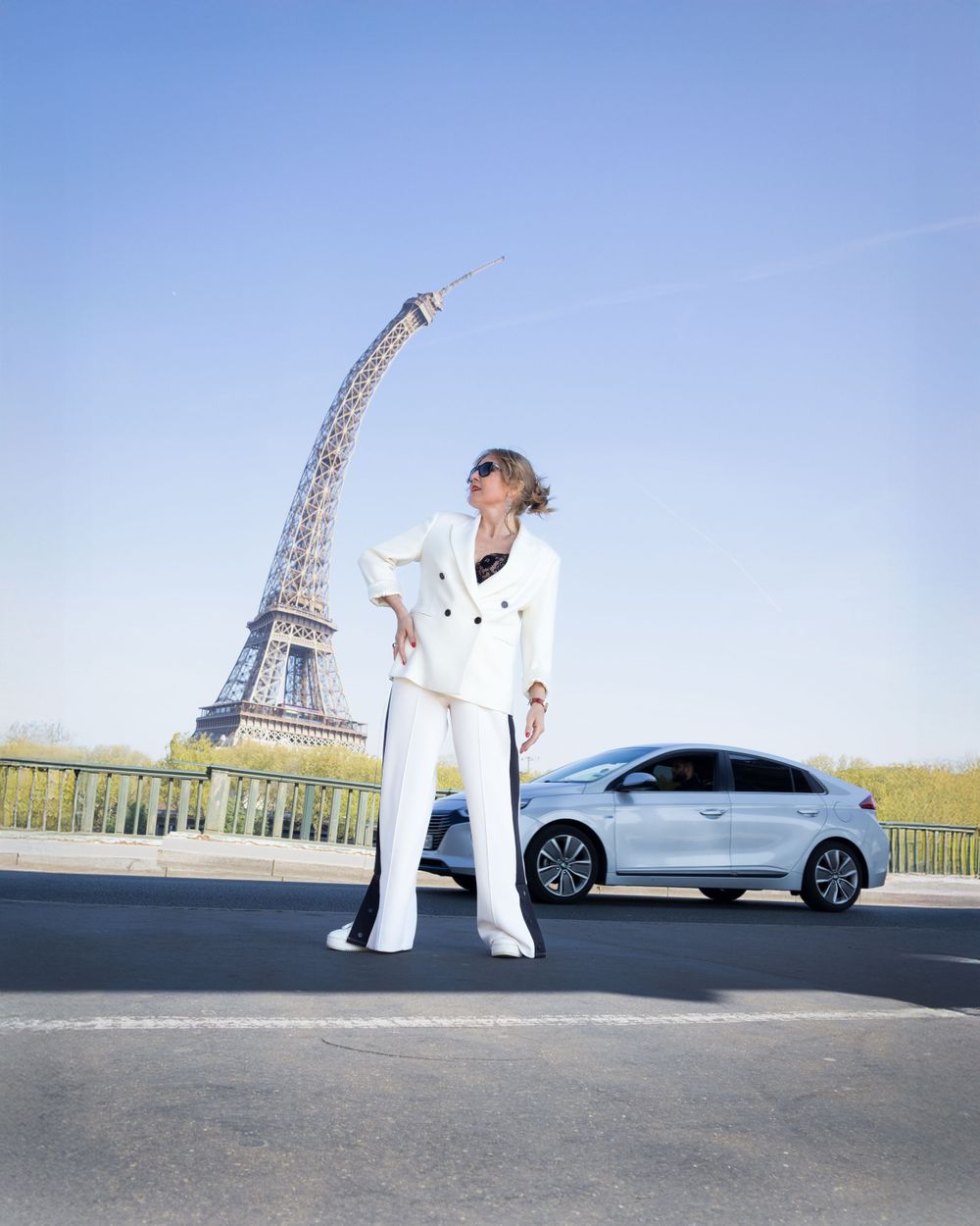 A person in a white outfit poses on a street with the Eiffel Tower and a silver car in the background.