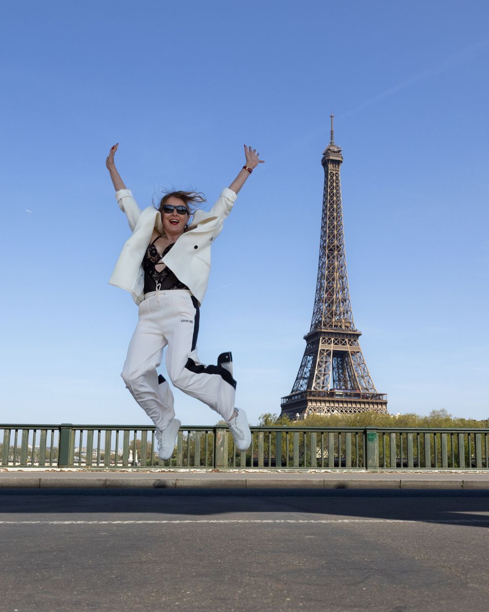 Someone in white attire jumps joyfully in front of the iconic Eiffel Tower in Paris on a sunny day.