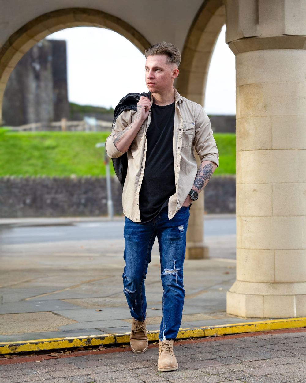 Casual street style portrait of model in beige jacket and jeans beneath stone archway.