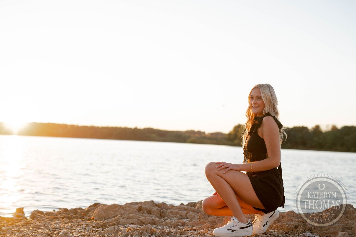 Sunset lakeside portrait of someone sitting on rocky shore in black outfit.