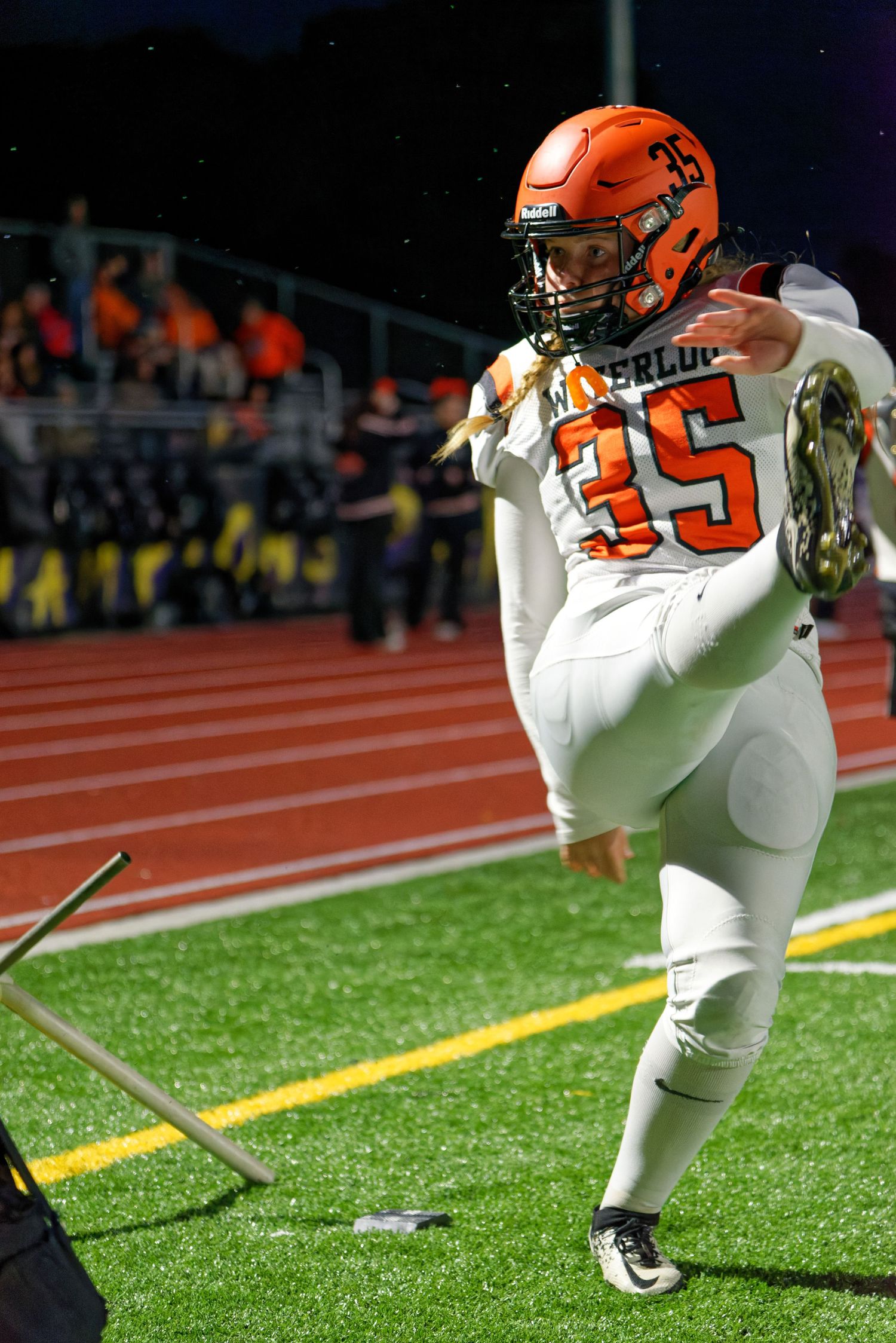 Female football player in orange and white warming up for field goal attempt during night game.