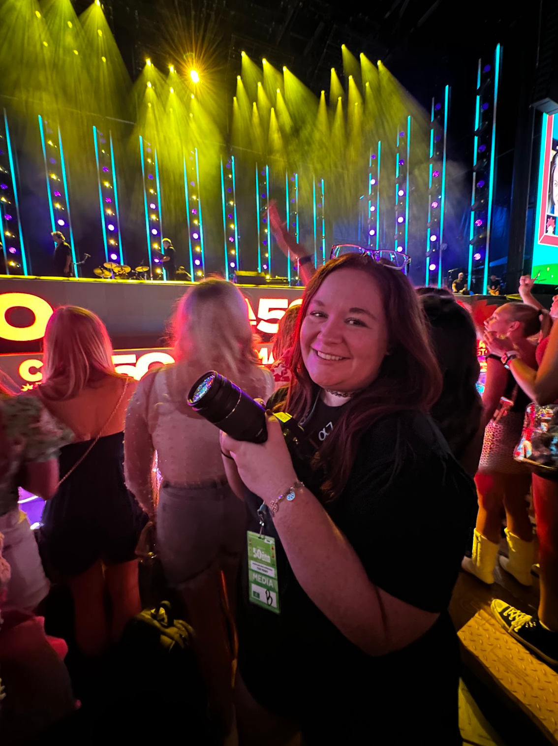 Concert crowd enjoying a nighttime performance with colorful yellow lighting and raised phones recording the show.
