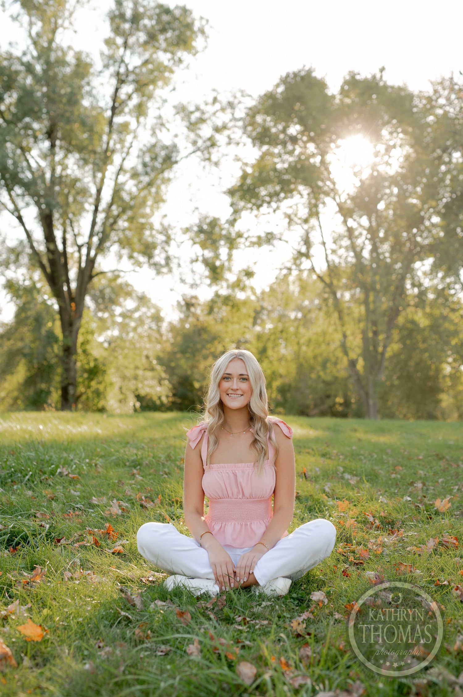 Outdoor portrait session with cross-legged pose on grass surrounded by fall foliage.