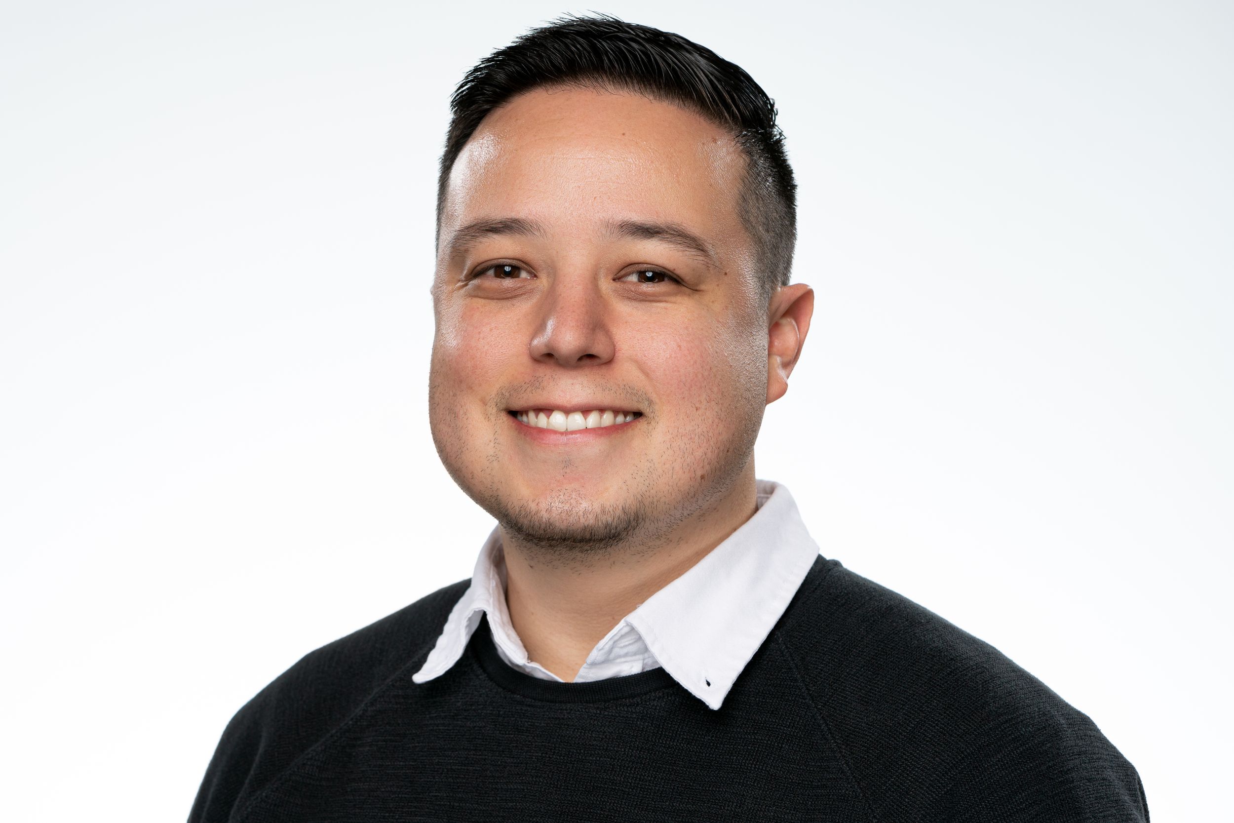 Professional headshot of person in black sweater with white collar smiling against light background.