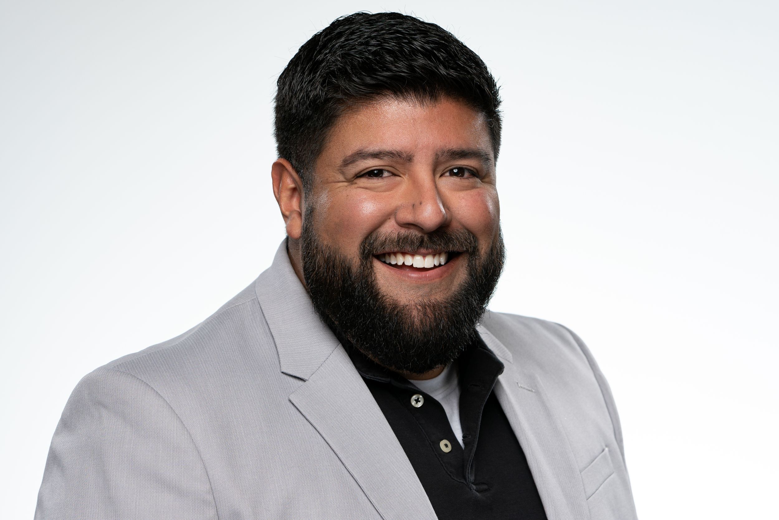 Professional headshot of person in gray blazer and black shirt smiling warmly against white background.
