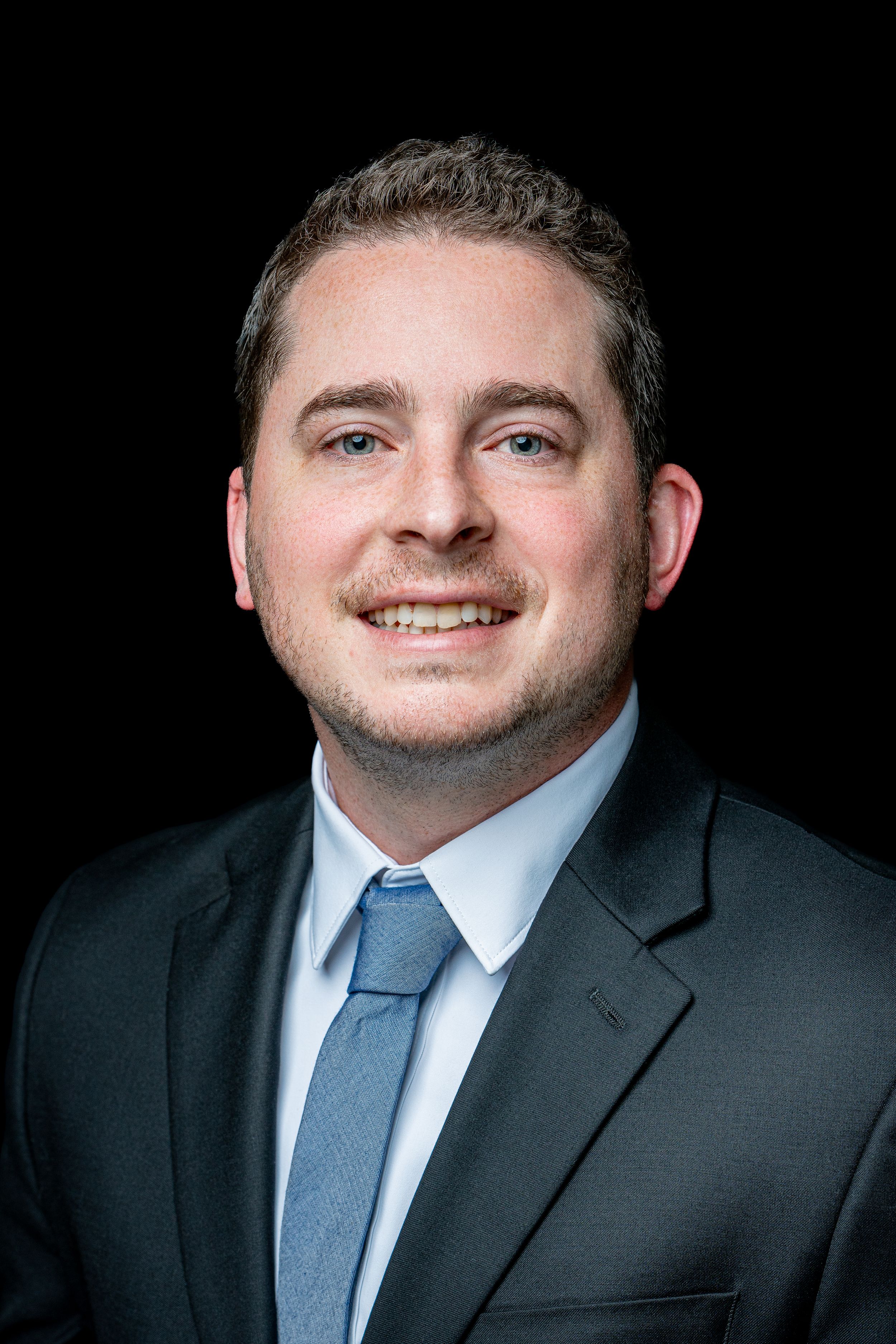 Professional business headshot of someone in a dark suit and blue tie against a black background.