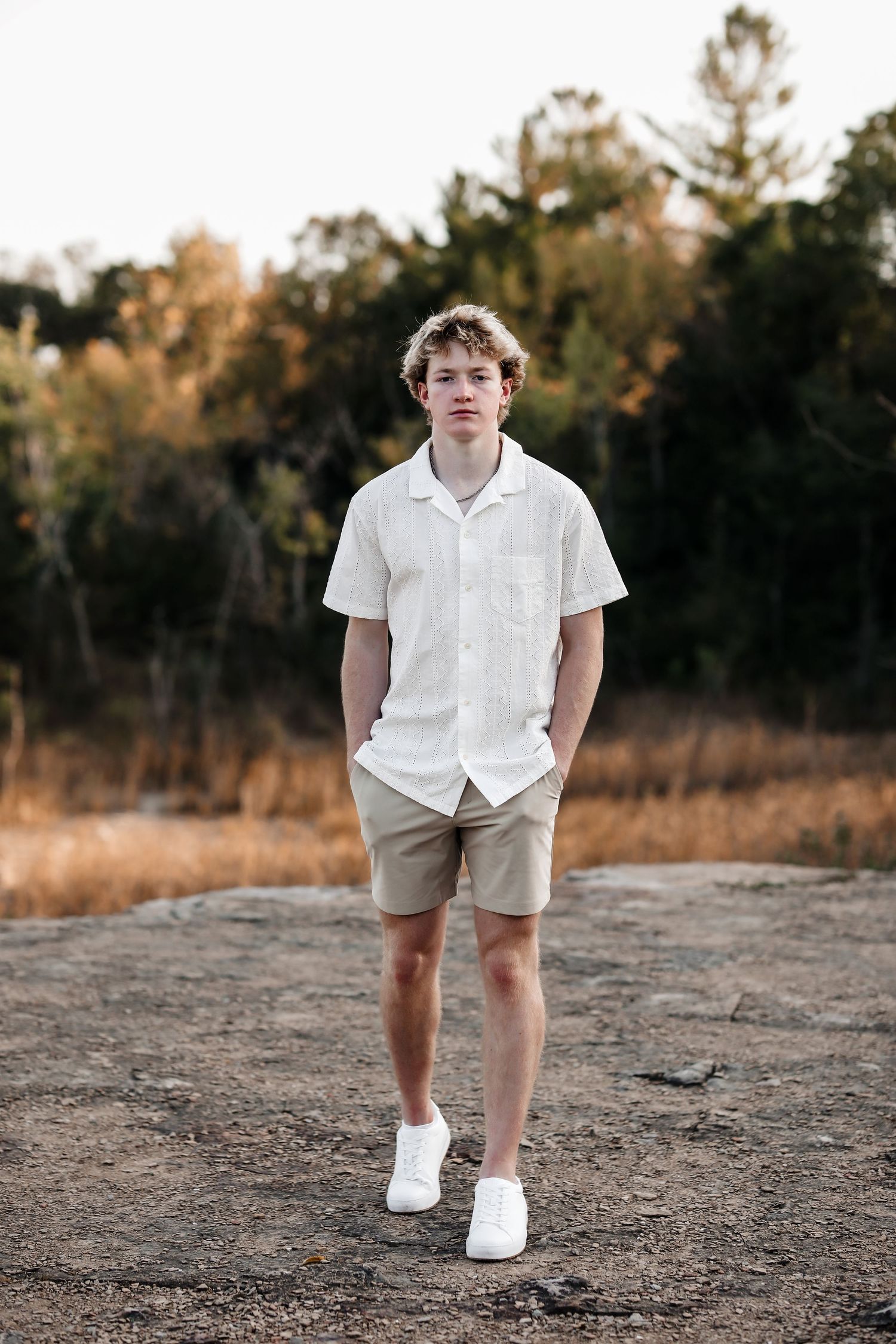 Teen boy in white shirt and khaki shorts standing on rock with autumn trees in background.