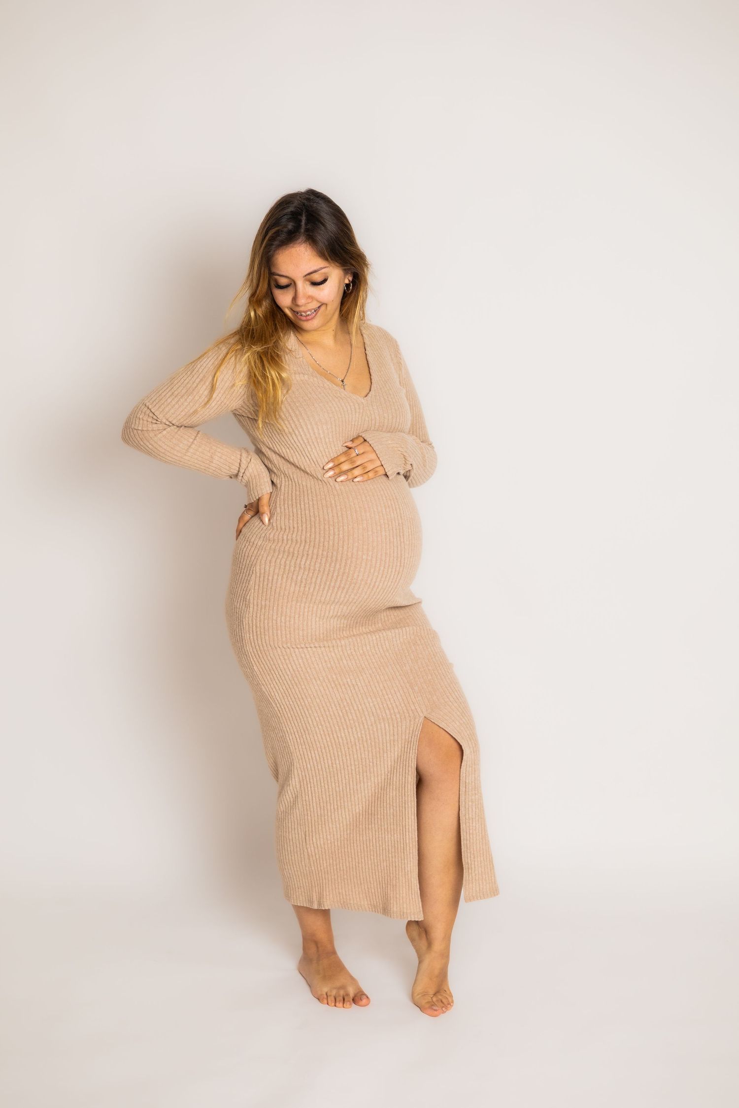 Maternity photoshoot with model in beige ribbed sweater dress on white background.