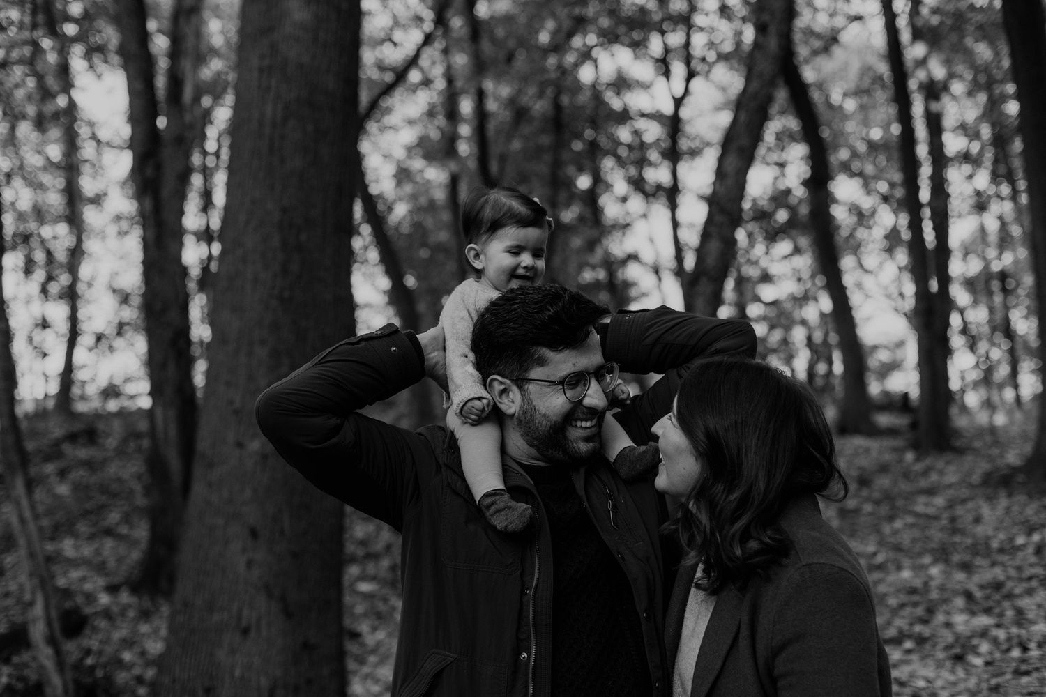 couple looking each other while he is carrying his 7 month baby on the shoulders, into the woods, in Toronto