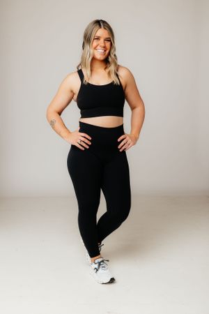 Woman in black athletic outfit including crop top and leggings stands confidently in studio against neutral backdrop.