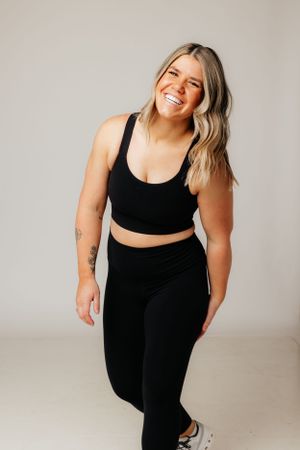Woman in black athletic wear sports bra and leggings smiling while posing against light gray studio background.