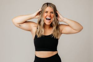 A joyful woman in black athletic wear laughs with hands raised to her head against a neutral background.