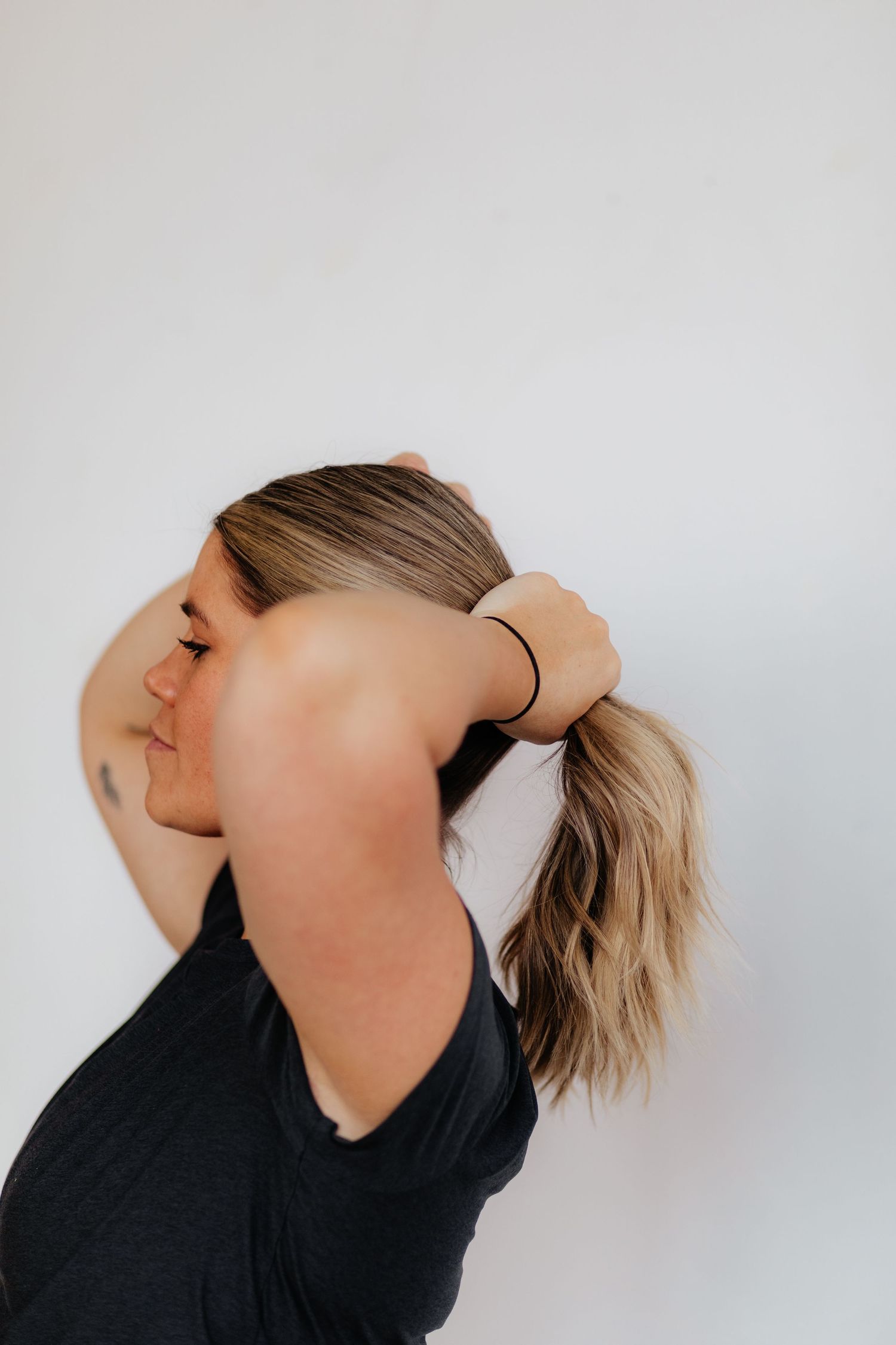 Person in black shirt adjusts their blonde ponytail against white wall background.