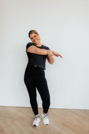 Athletic person in all black workout attire stretches arms while exercising in a bright studio space.