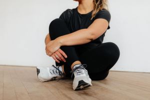 Person in athleisure attire sitting cross-legged on wooden floor wearing gray sneakers and black workout clothes.