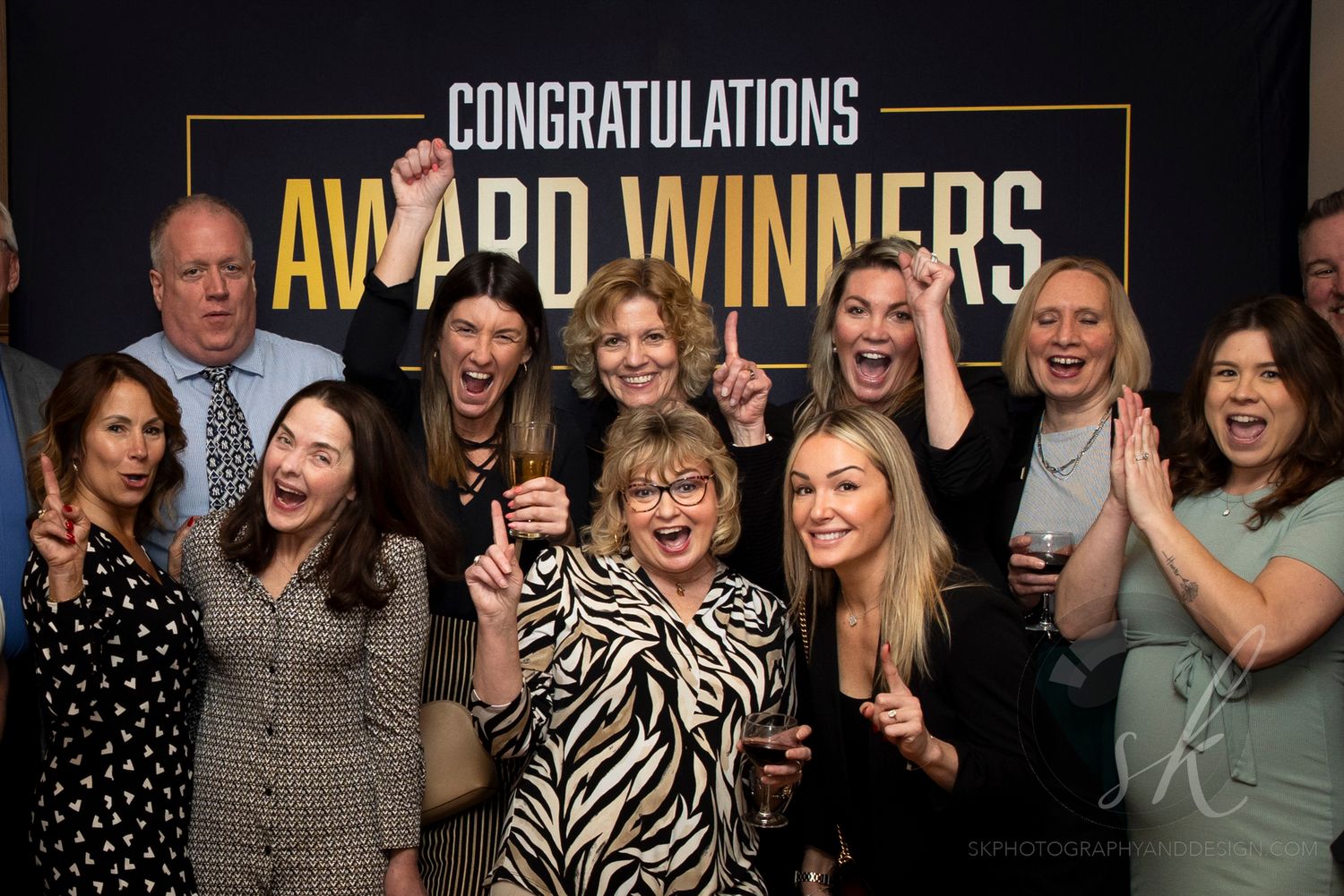 Group of award winners celebrating in front of a Congratulations Award Winners sign, cheering and smiling.