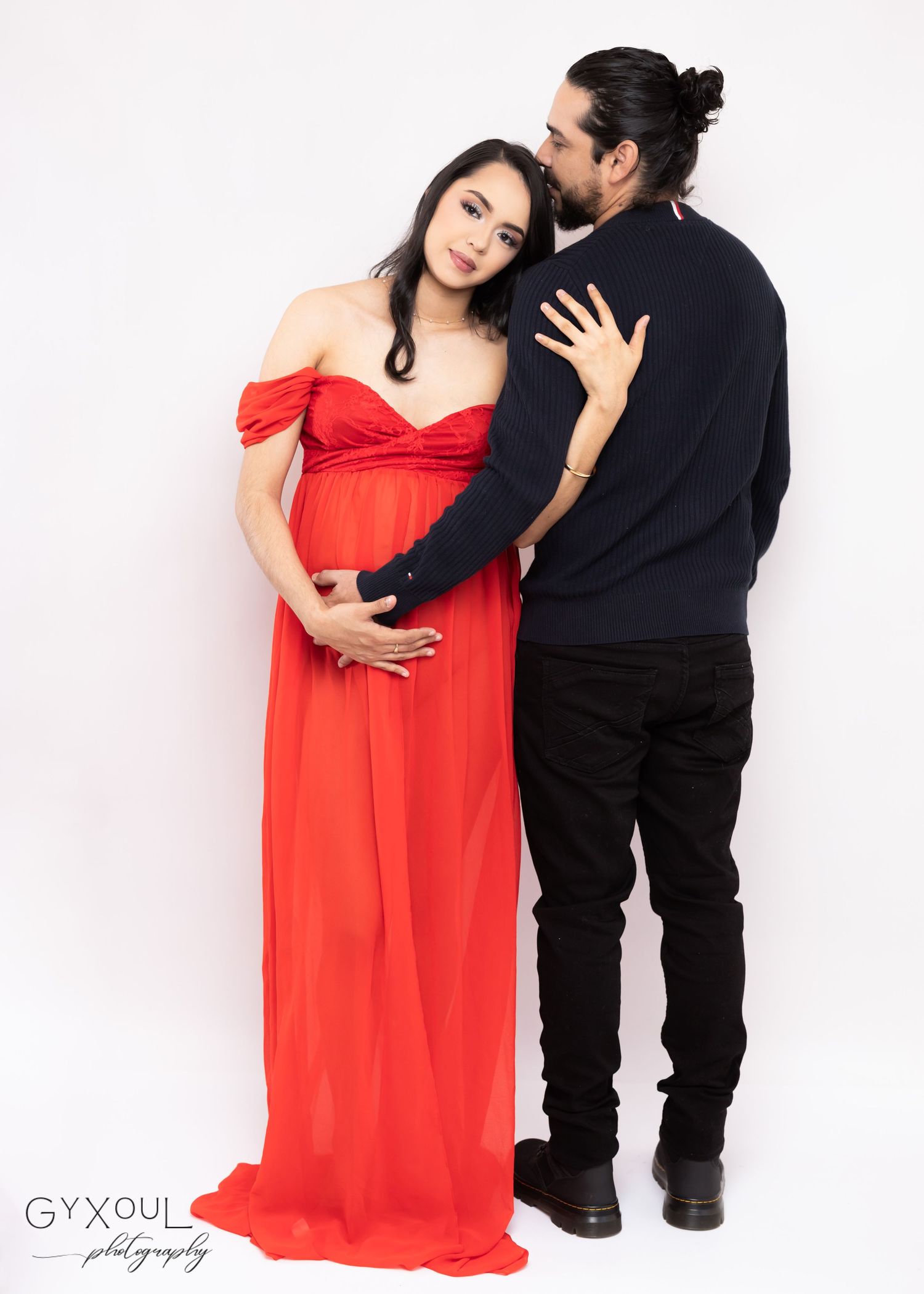Romantic maternity photo with couple in red dress and black sweater against white background.