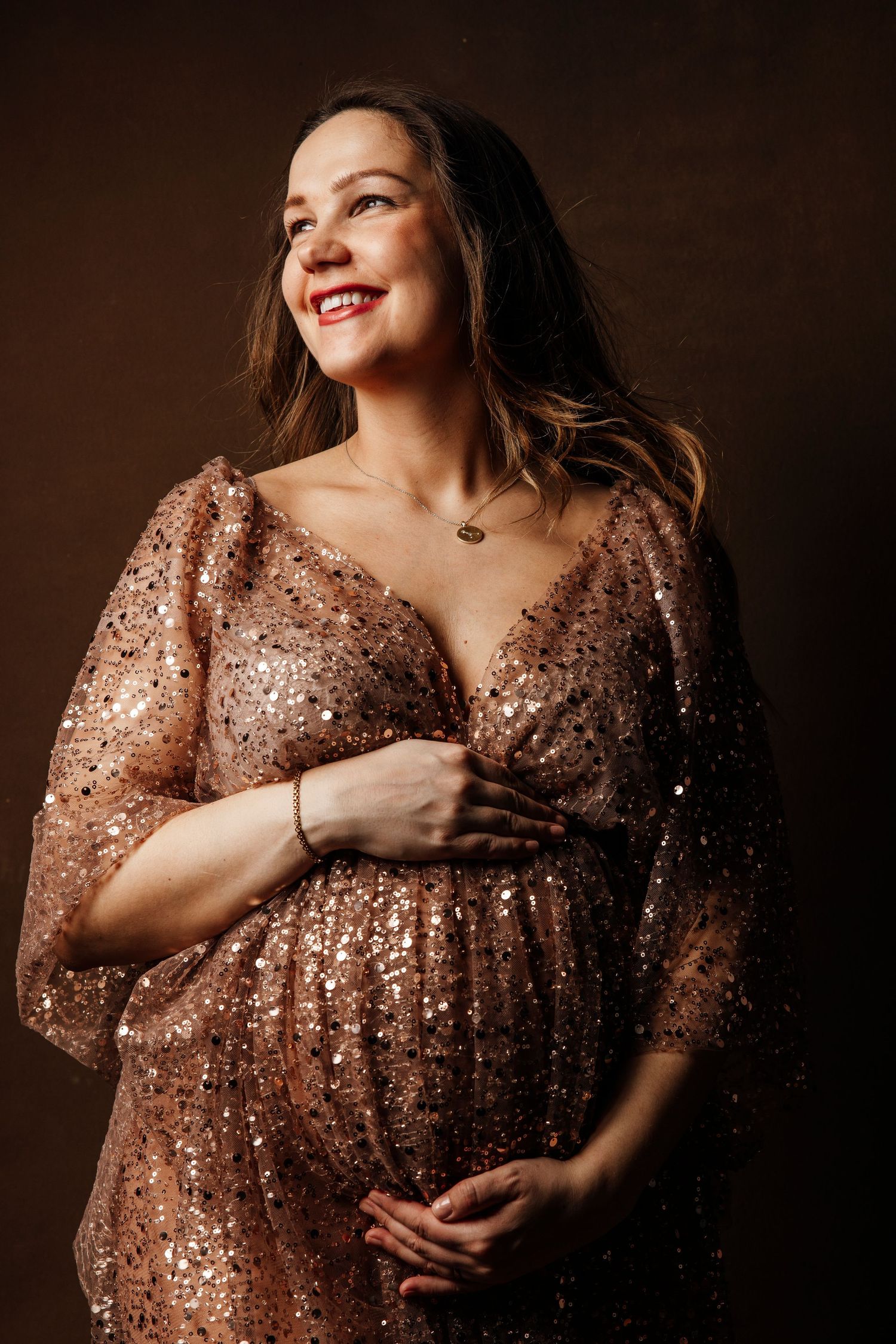 Expectant mother in sparkling rose gold dress poses against dark backdrop.
