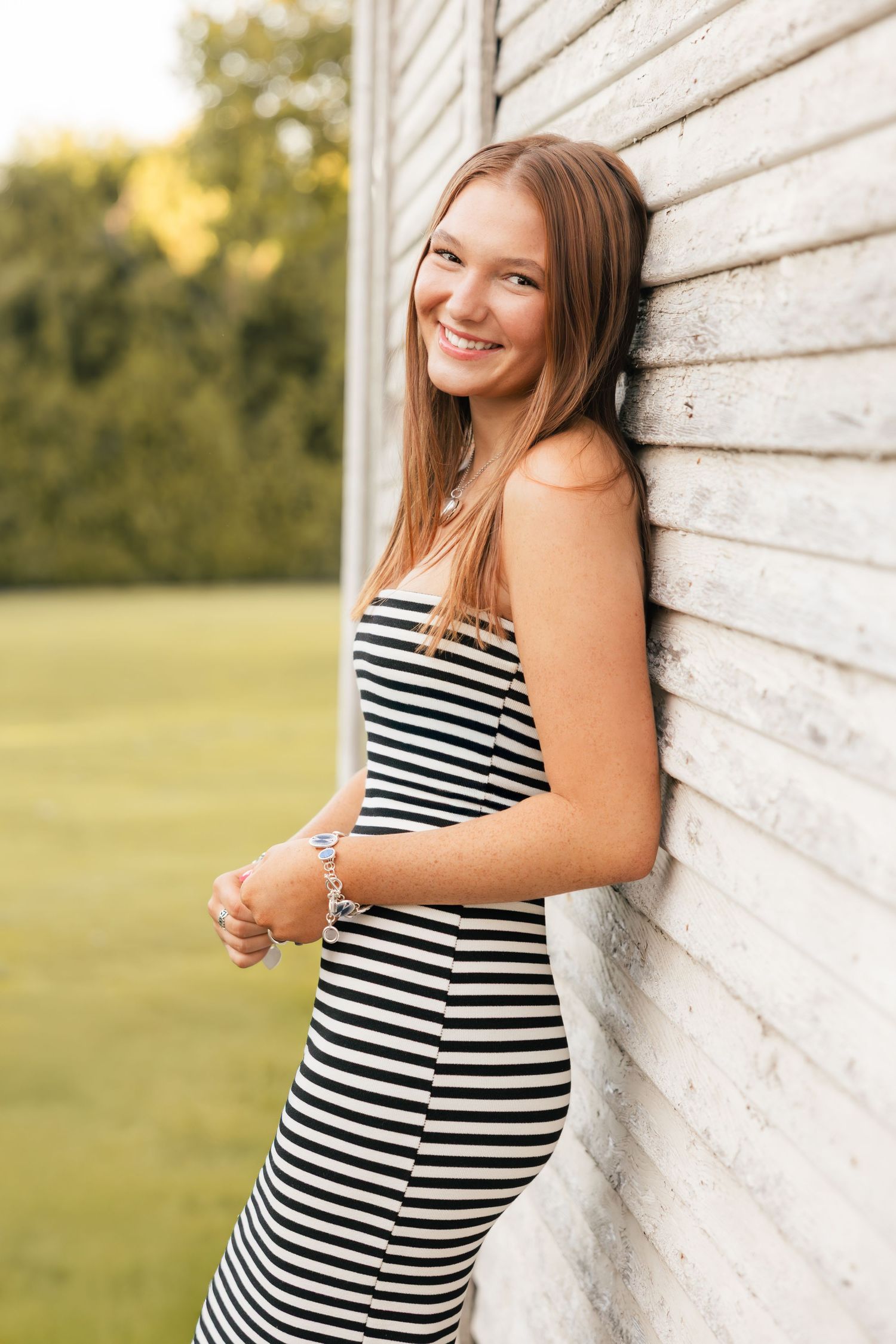 A pregnant person in a striped dress poses against a rustic white wall during a maternity photoshoot in golden sunlight.