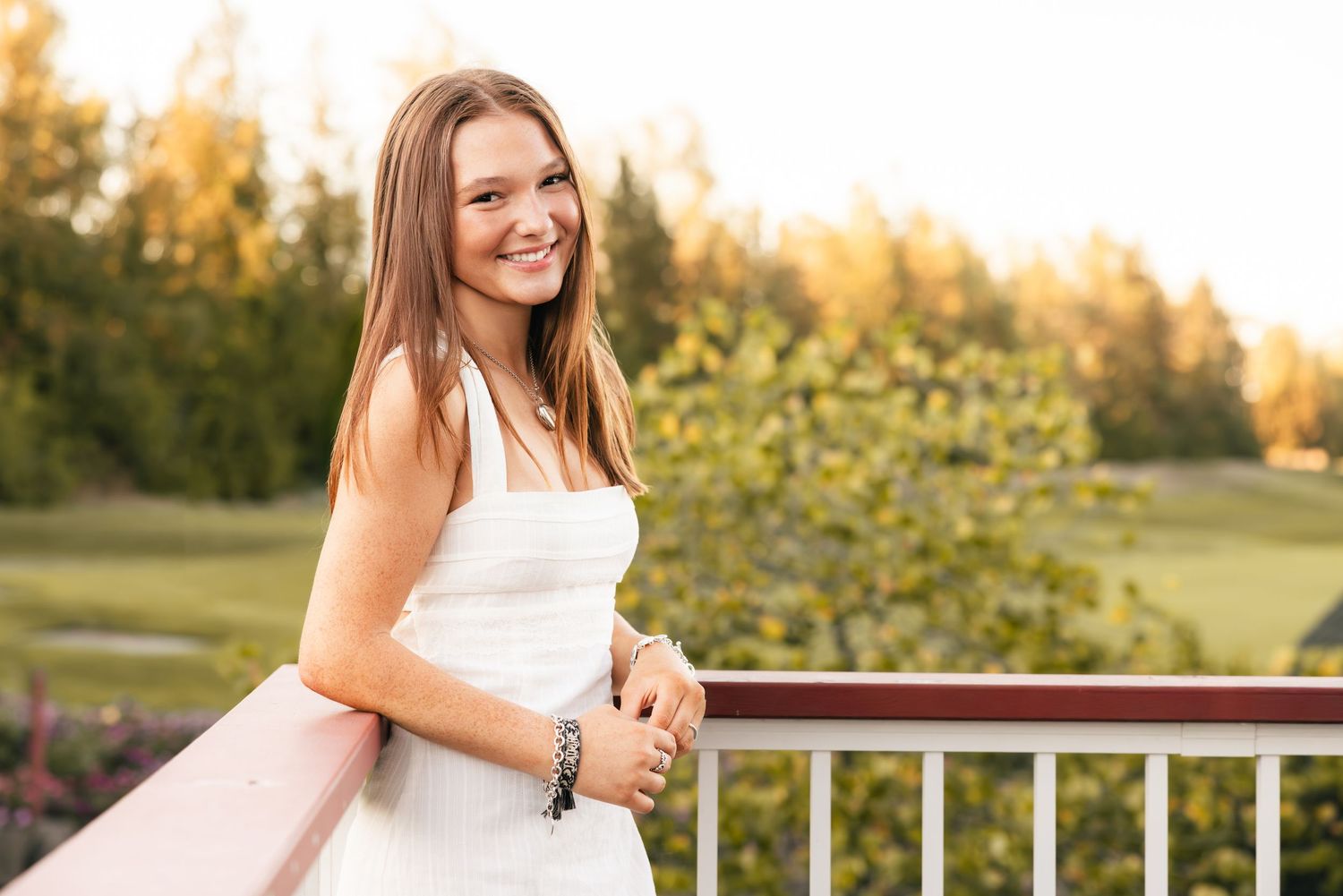 A person in a white dress poses joyfully on a wooden deck at golden hour in a scenic outdoor setting.