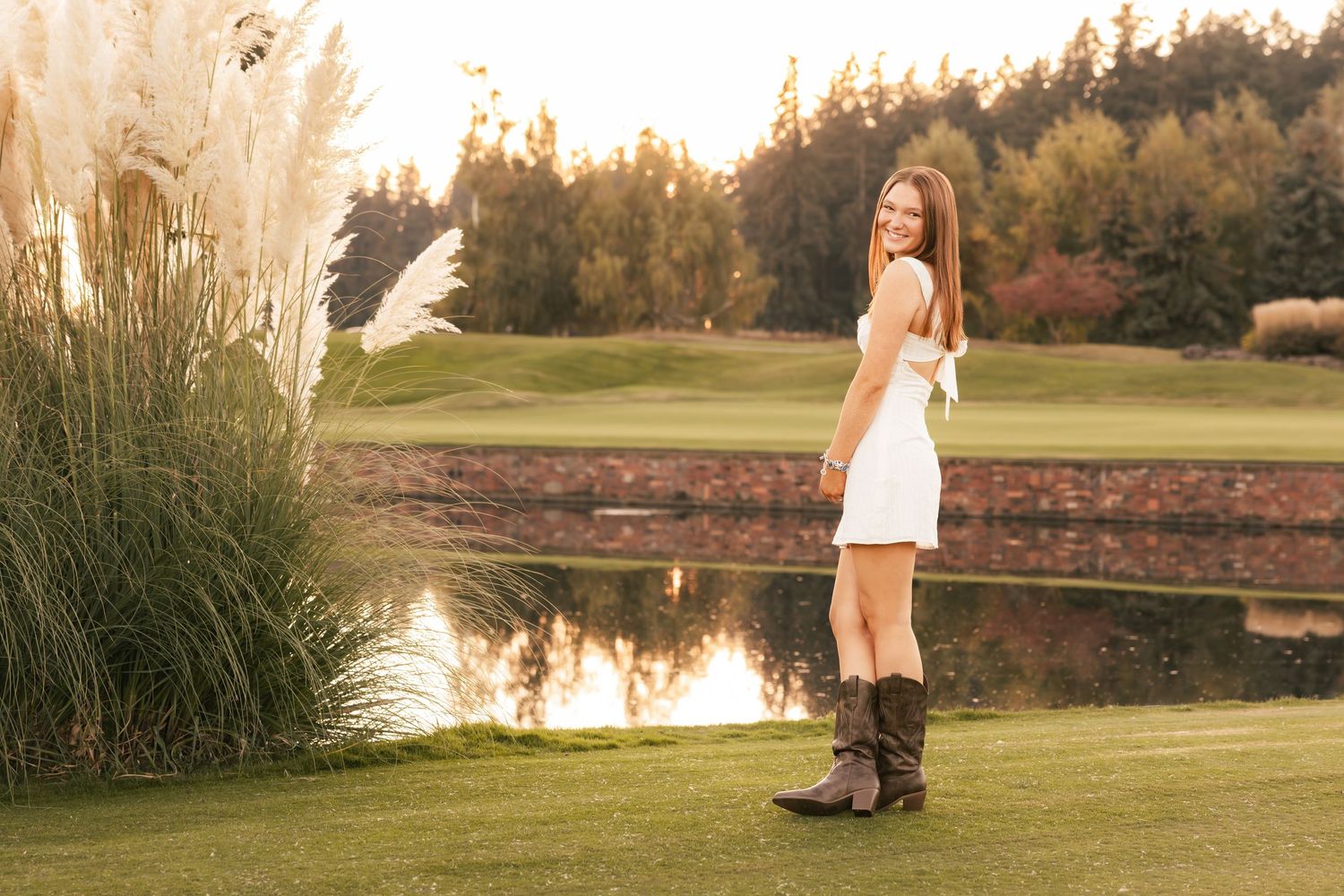 Someone in a white dress and cowboy boots stands on a golf course at sunset near tall ornamental grasses and a pond.