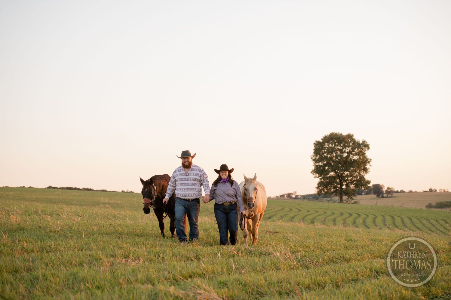 Couple walking with two horses up a grassy hill during golden hour.