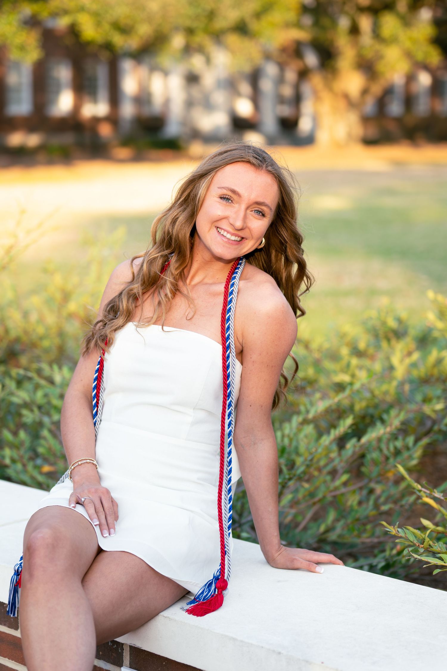 A person wearing a white dress sits outdoors on a bench with braided red and blue cords in warm golden sunlight.