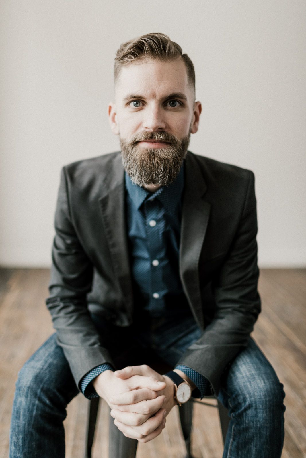 Professional portrait of a bearded person in a dark blazer and blue shirt sitting casually on a wooden floor | Photographer Josh Ohms