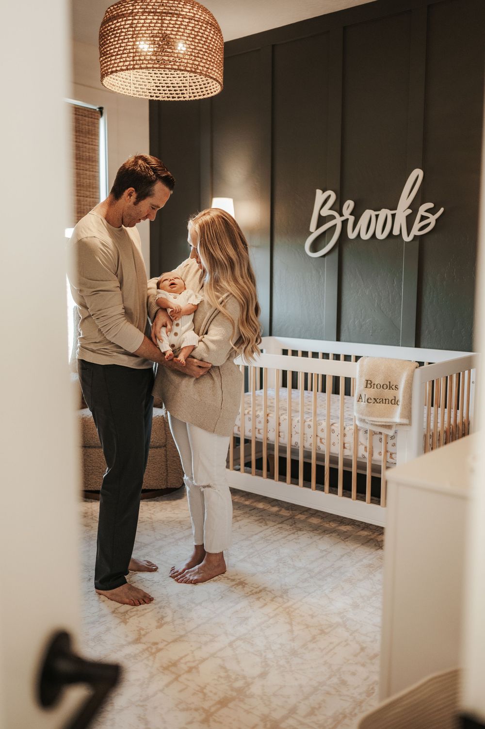 Cozy nursery featuring woven pendant lamp, dark accent wall, and white crib with name sign.